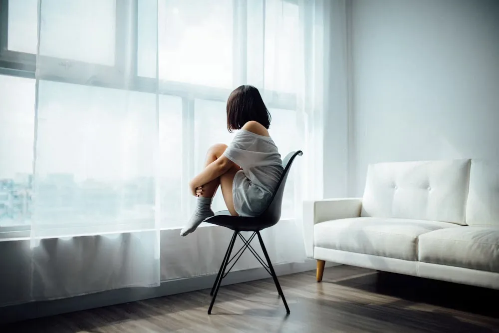 girl with shoulder-cut black hair wide neck white t-shirt sitting on a black table with raised legs and arms across like hug facing window having a white translucent curtain and a white sofa in diagonal direction