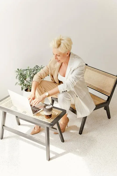 a woman with blonde hair working on a laptop wearing half nude color and half white color blazer and pant with light brown high heels