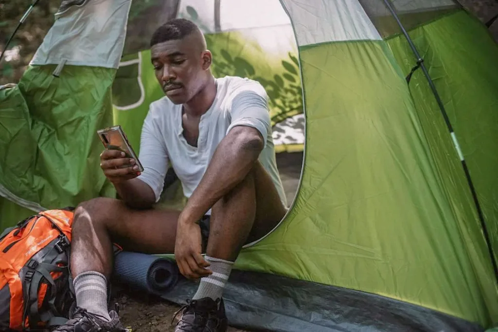man sitting in a tent in white t shirt holding phone in right hand and seeing it - Online Dating Profiles
