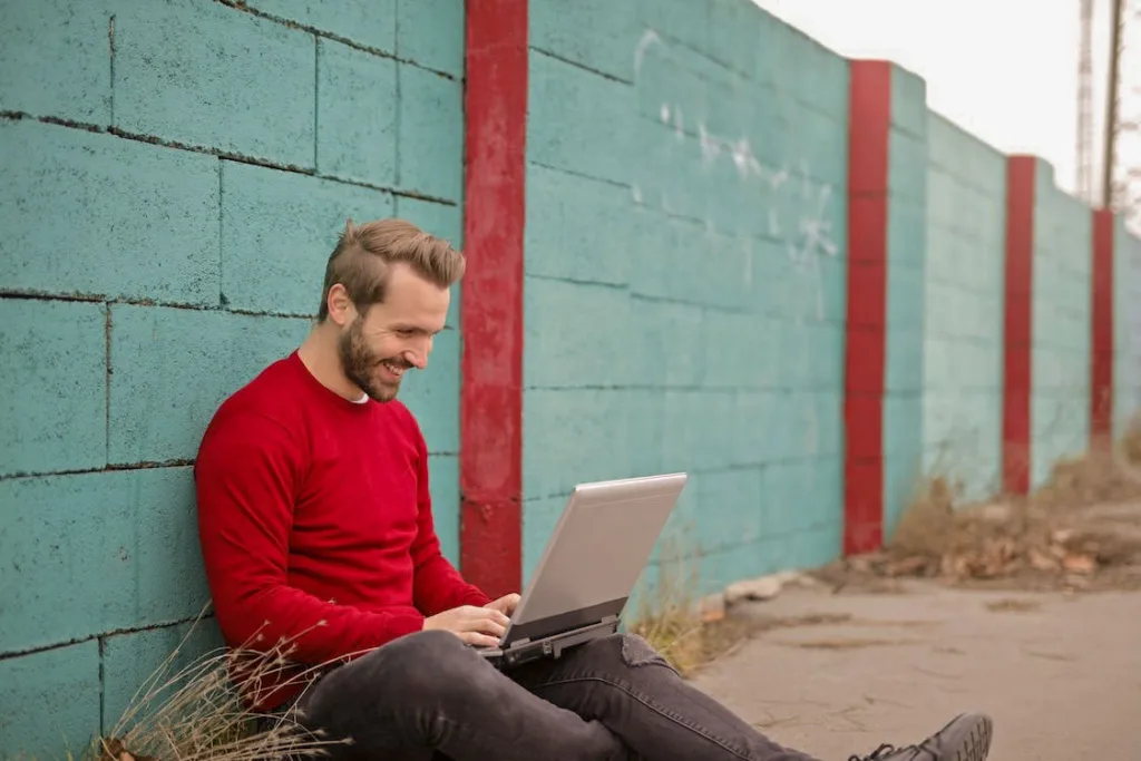 man in red round neck full sleeves shirt leaned against the wall and smiling on laptop - Online Dating Success