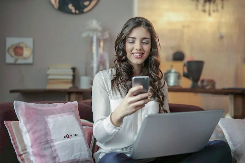 woman with laptop on lap holding mobile phone in right hand and smiling - Online Dating Success