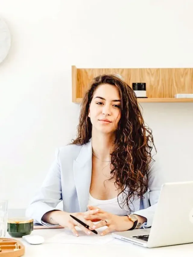 woman sitting in front of laptop on her desk with open hair in sky blue coat and white round neck t shirt