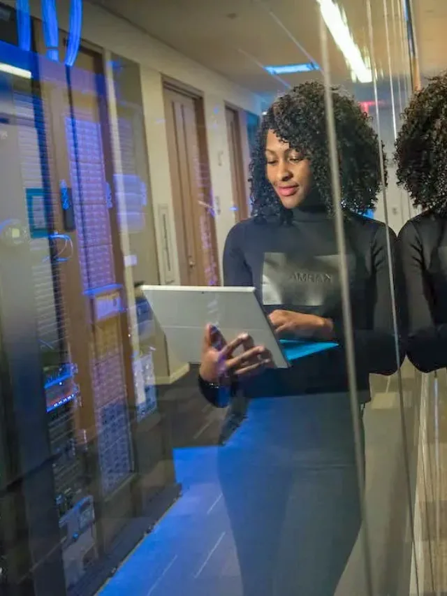 woman in black full neck full sleeves and black pant leaning against a glass wall holding laptop in hand and typing in it.