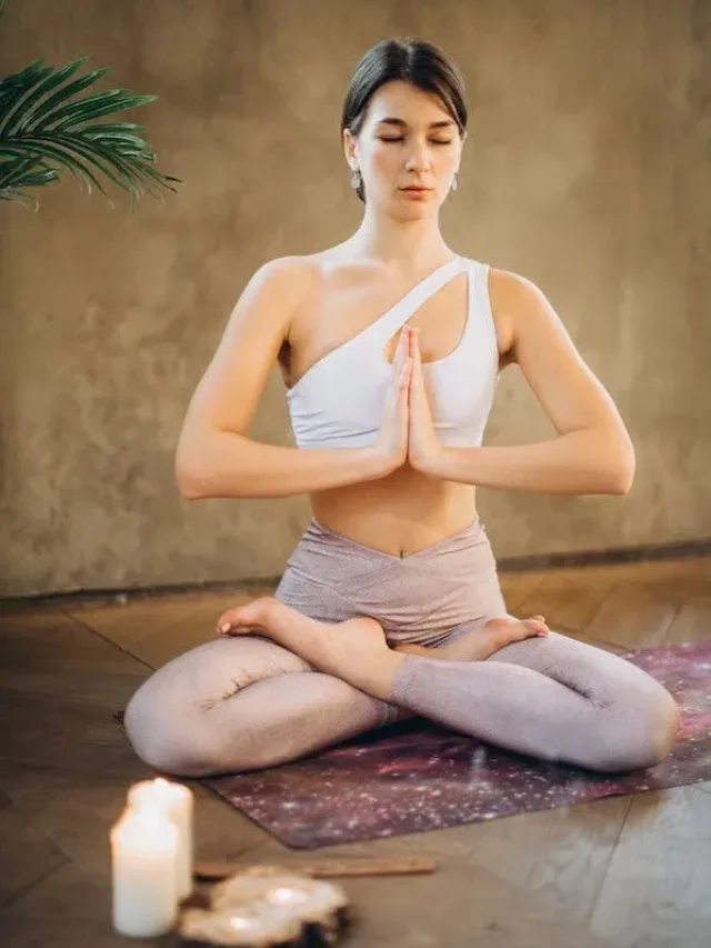 woman in padmasan posture with folded hand in sports bra & yoga pants and a lighted scented candle