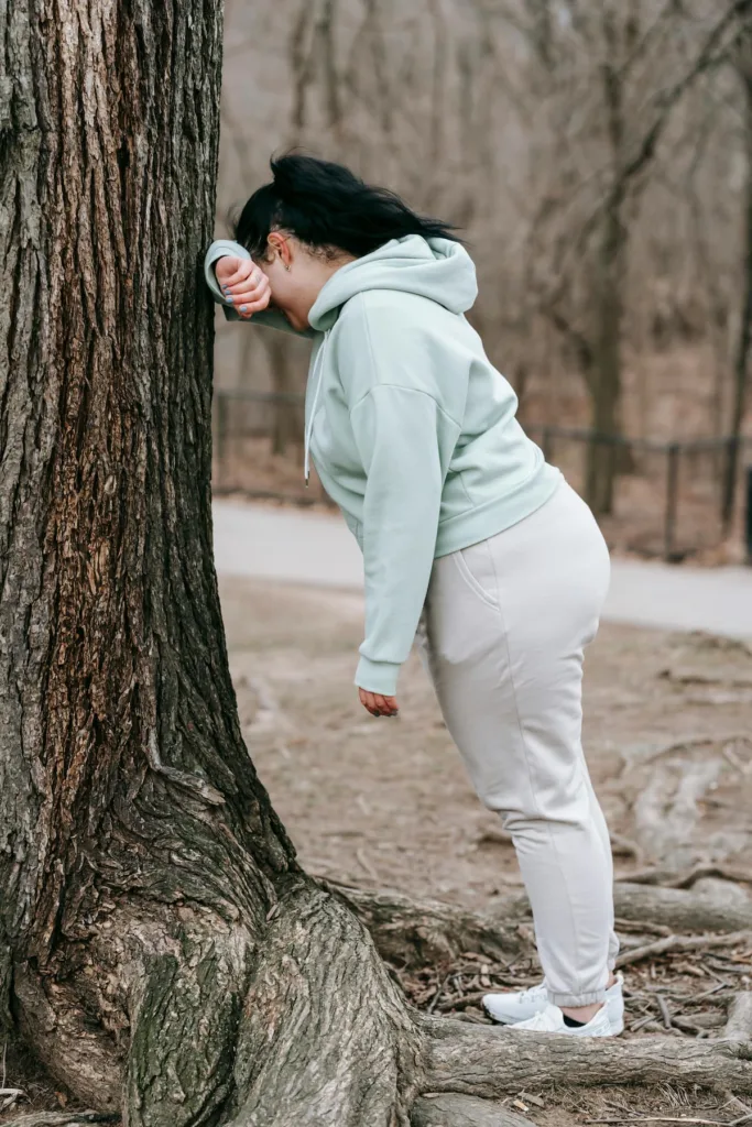 woman in woods with aqua hoodie and white pants with left hand on head and leaning against the tree showing ADHD's Impact on Women's Mental Health