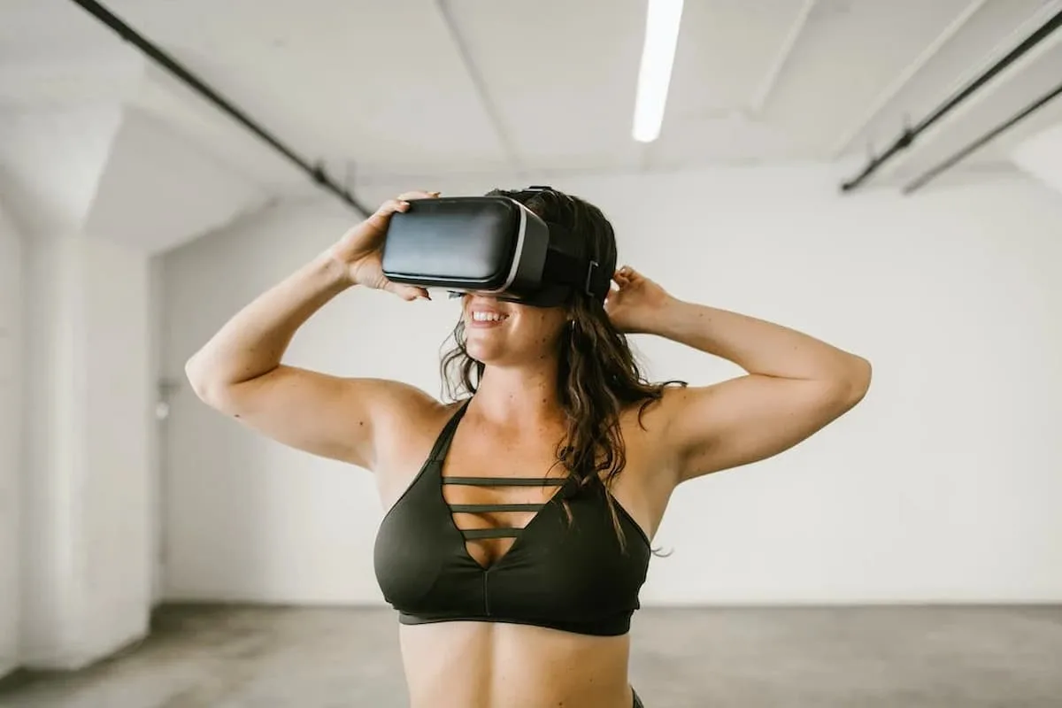 Woman earing VR headset and black sports bra smiling inside a hall like place.