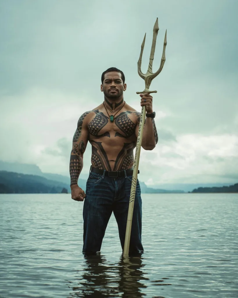 man with ribbed body with tattoo standing in water holding trident of aquaman