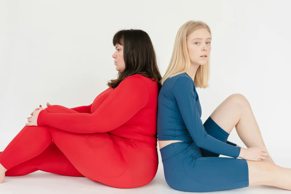 one healthy woman in red and other slim woman in aqua blue sitting with back touching each other
