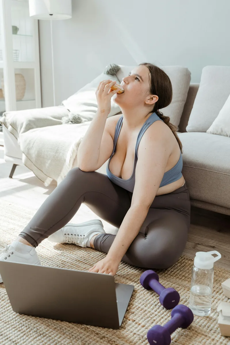 A healthy woman eating burger with right hand looking up and typing on a laptop besides dumbles and a jar of water -Rapid Weight Loss
