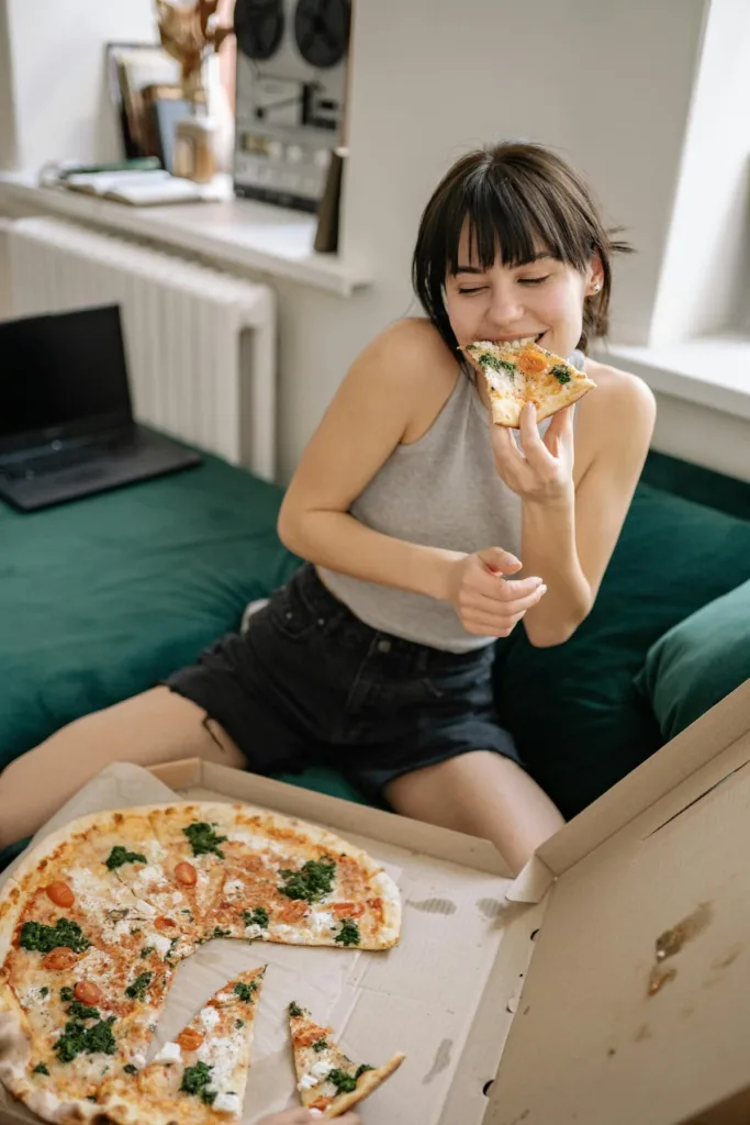 woman in halter neck gray top with dark grey denim shorts eating a slice of pizza with left hand couching on a green sofa.