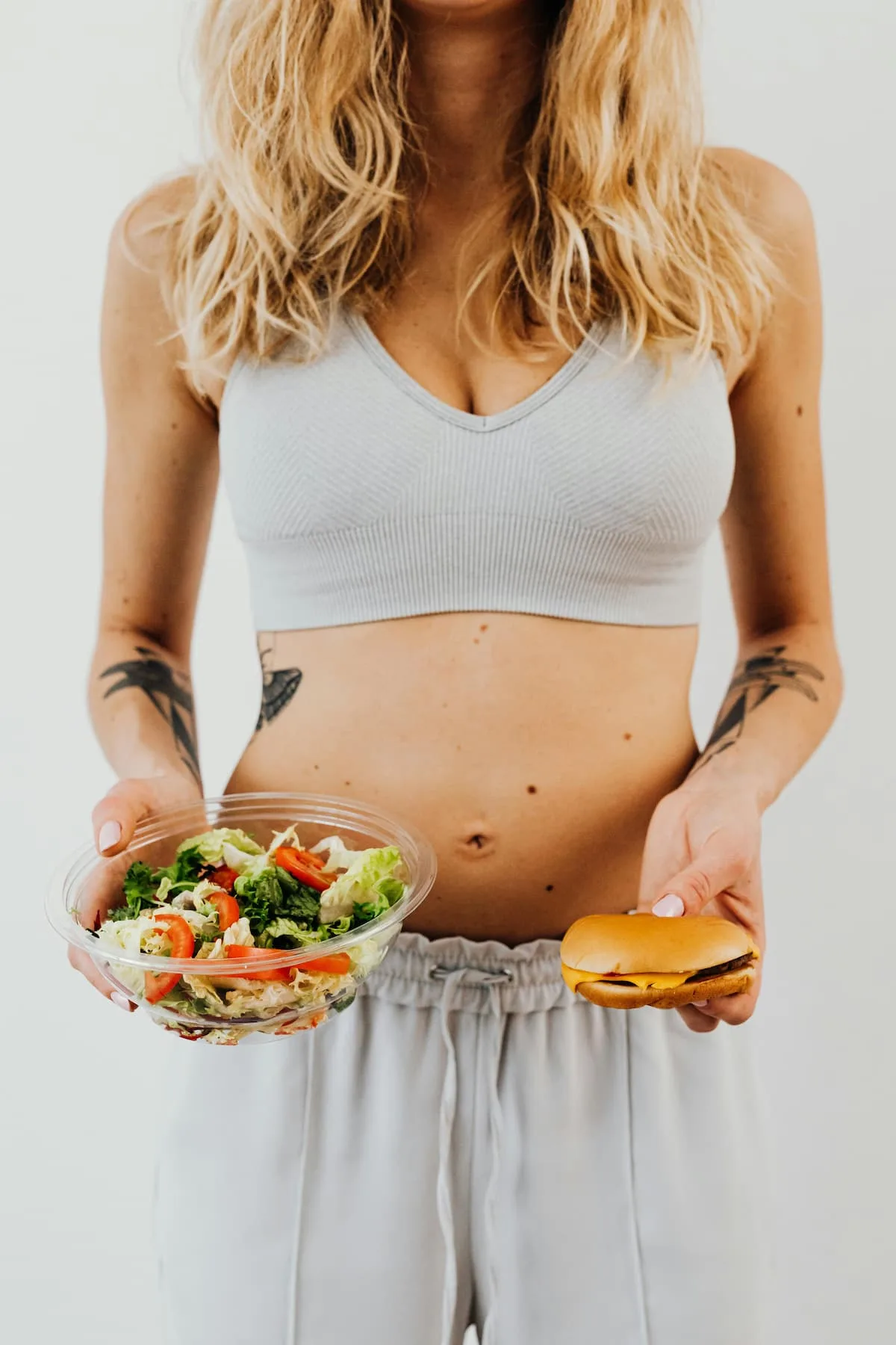 woman in sports gray bra and night pants holding salad bowl in one hand and burger in other hand Ultra Processed Foods and Weight Gain