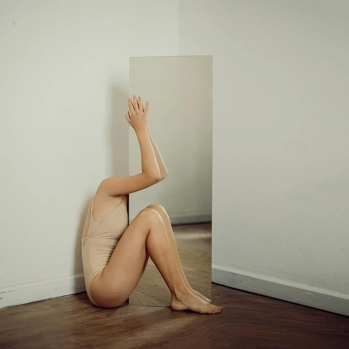woman sitting in nude body suit with a face hiding against the mirror and reflection of one hand and leg seen in mirror