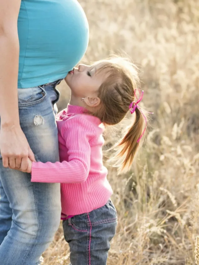 pregnant woman in blue top being kissed on belly by a small girl in pink top