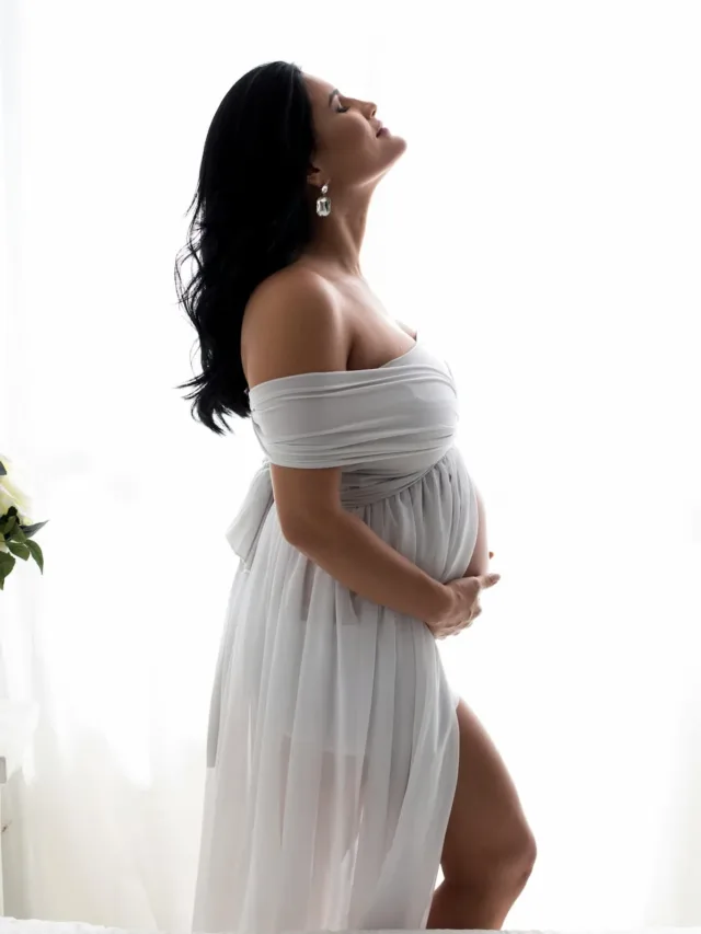pregnant woman doing a photo shoot in white gown closed eyes looking in sky and holding the belly with one thigh high slit