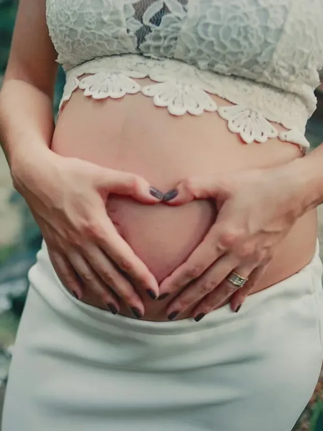A pregnant woman in white top making a heart shape on her belly with both hands