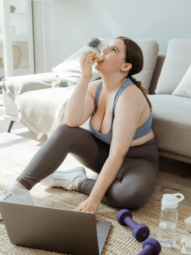 A healthy woman eating burger with right hand looking up and typing on a laptop besides dumbles and a jar of water -Rapid Weight Loss