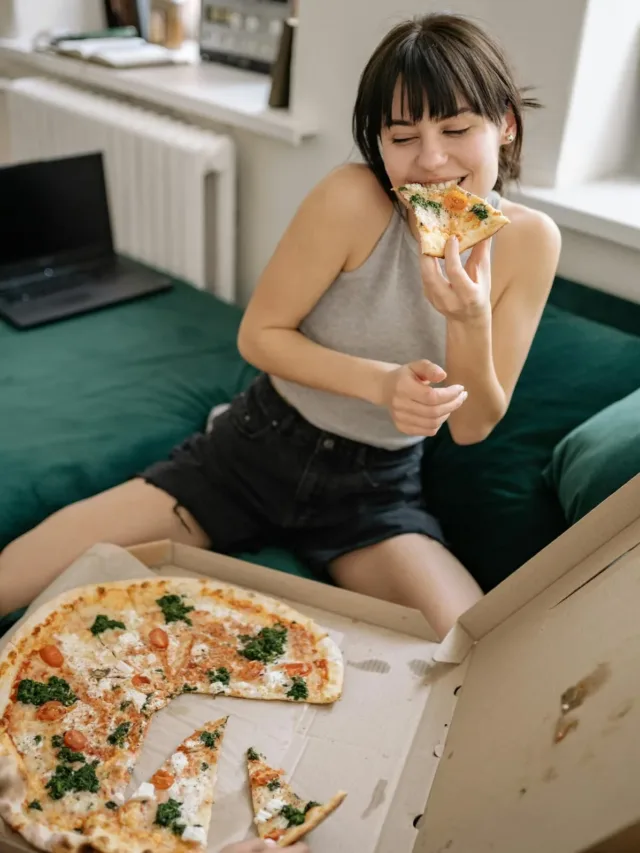 woman in halter neck gray top with dark grey denim shorts eating a slice of pizza with left hand couching on a green sofa.